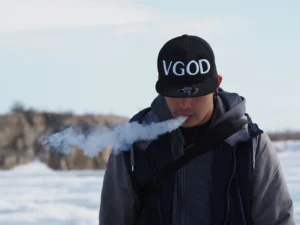 一位戴著「VGOD」品牌帽子和灰色連帽衫的成年男性老玩家，在模糊的雪地戶外冬日場景中專注地吞吐厚實的白色電子煙霧化蒸汽。這是一張用於現代替煙方案指南的特色圖片。